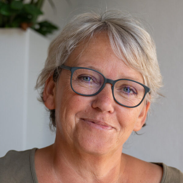 Porträt einer lächelnden Frau mit kurzen grauen Haaren und Brille, aufgenommen im natürlichen Licht vor einem hellen Hintergrund.